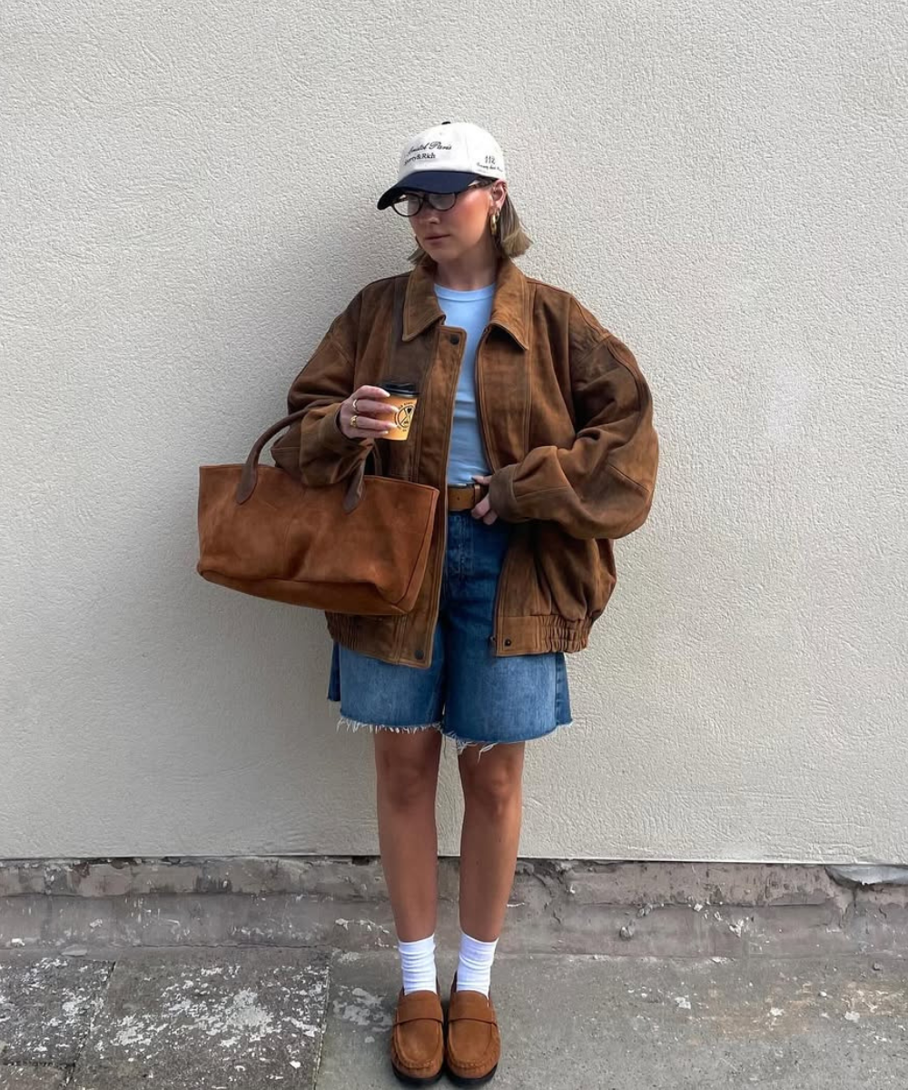 Pessoa vestindo jaqueta marrom com short jeans, boné branco e bolsa marrom. Usa sapatos combinando.