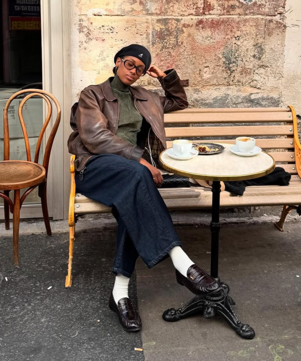 Pessoa sentada em banco, vestindo jaqueta marrom com calça escura, beret preto e sapatos marrons, ao lado de mesa com xícaras.