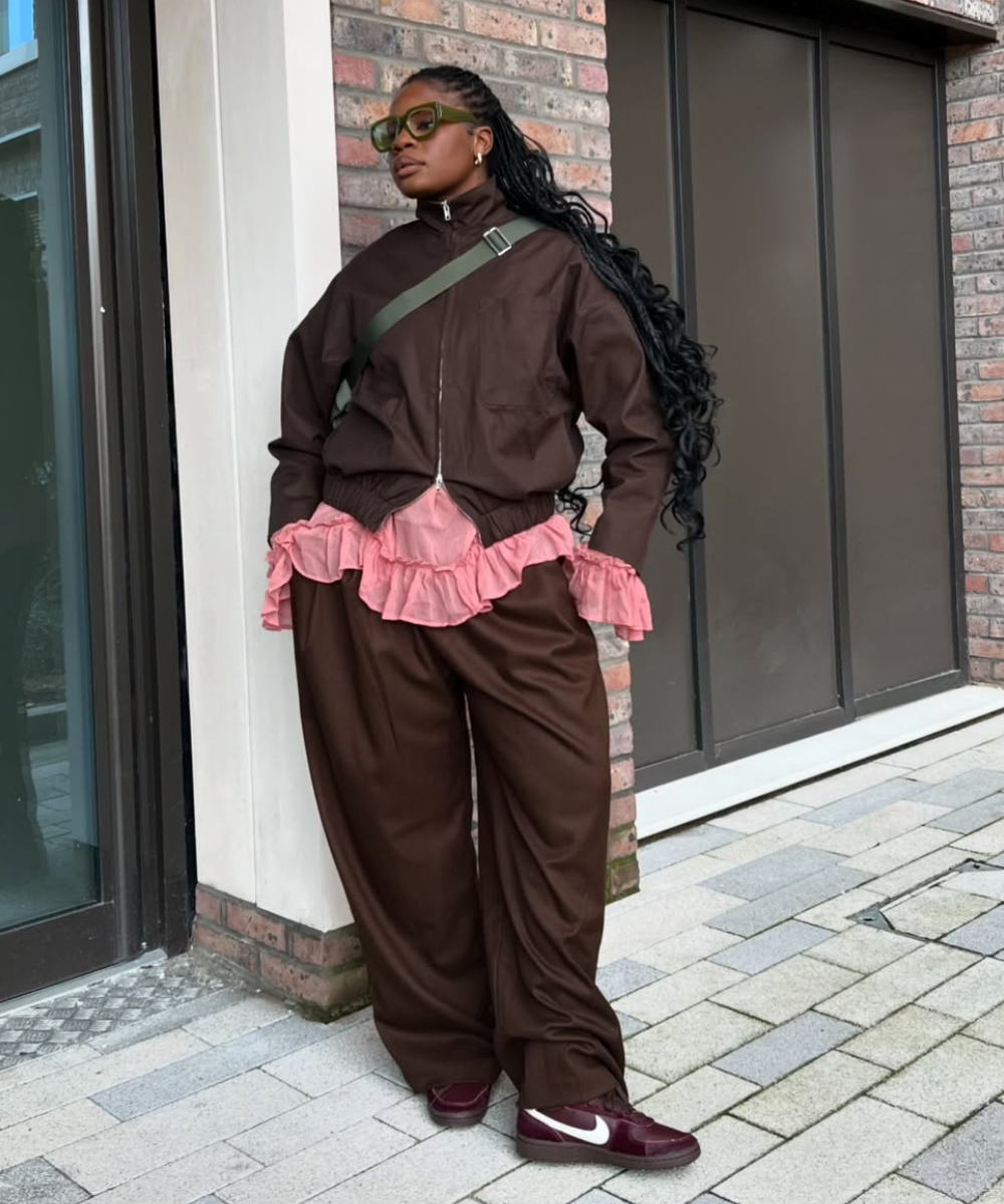 Pessoa veste jaqueta marrom sobre blusa rosa com babados, calça marrom e sapatos esportivos.