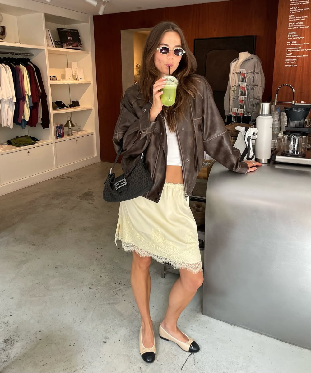 Pessoa com jaqueta marrom, óculos escuros, segurando uma bebida verde em loja de roupas elegante. Mostra como usar jaqueta marrom.