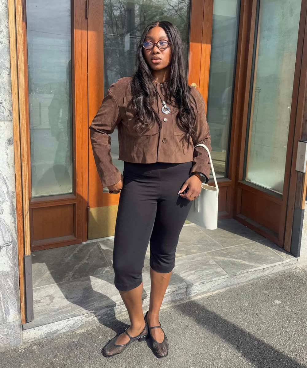 Pessoa em frente a porta veste jaqueta marrom e calça preta, usando óculos de sol.