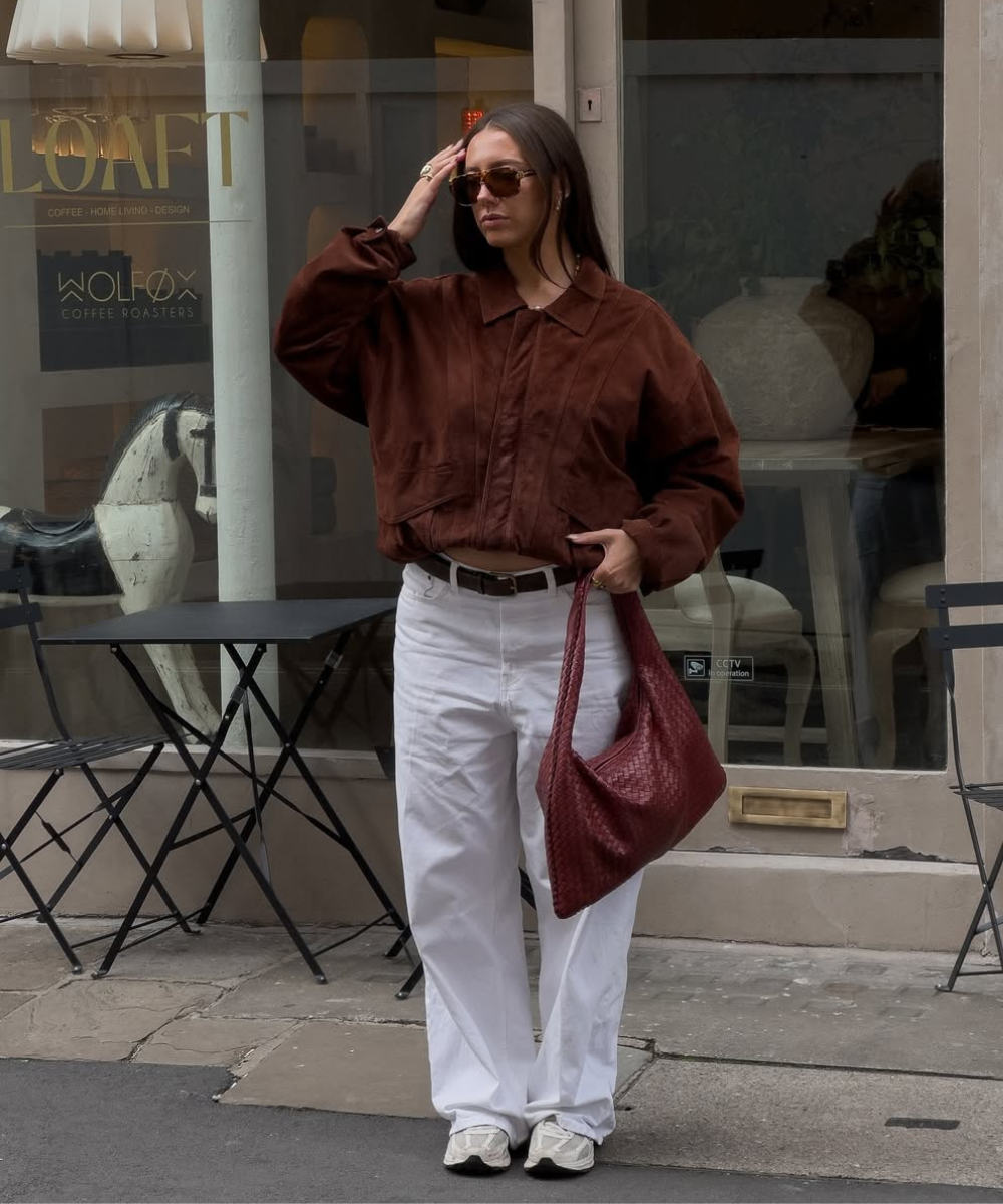Pessoa usando jaqueta marrom combina com calças brancas e bolsa vermelha em frente a um café.