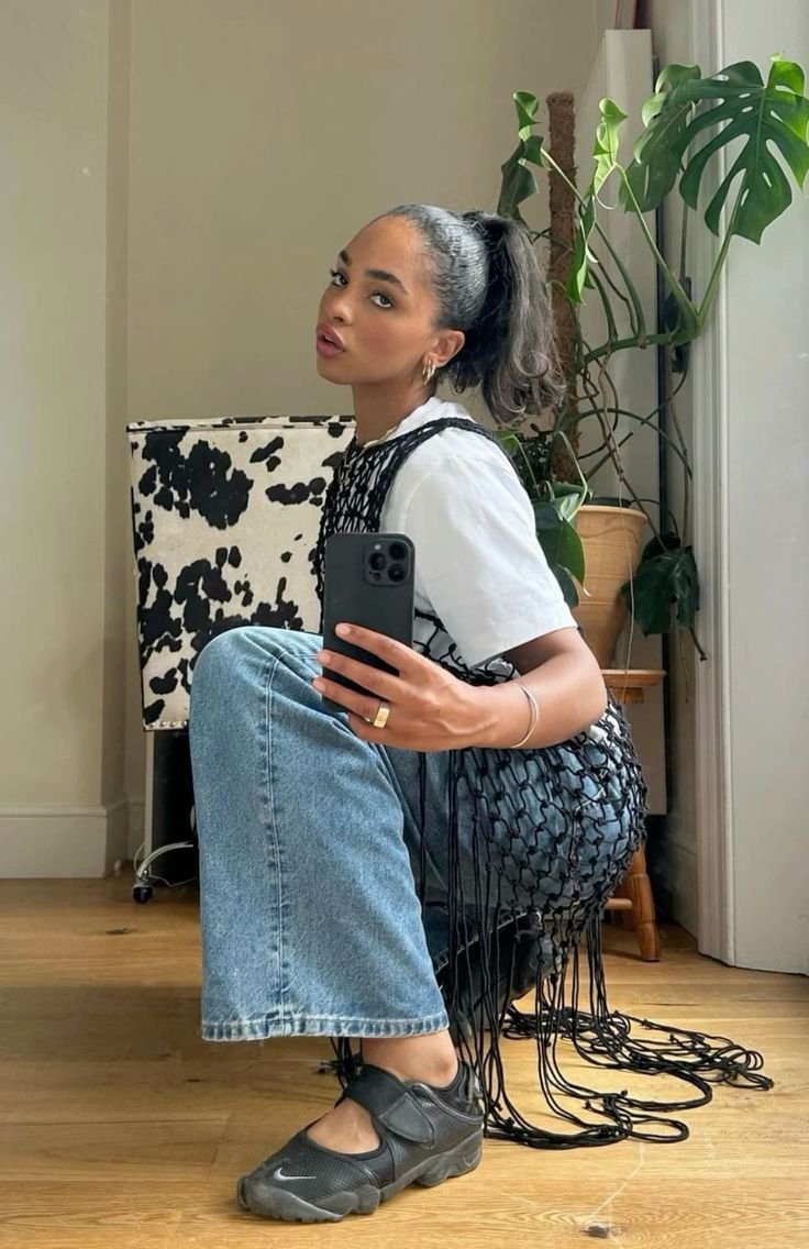 Mulher sentada de perfil, com camiseta branca e calça jeans, usando Nike Air Rift pretos, tirando foto no espelho.