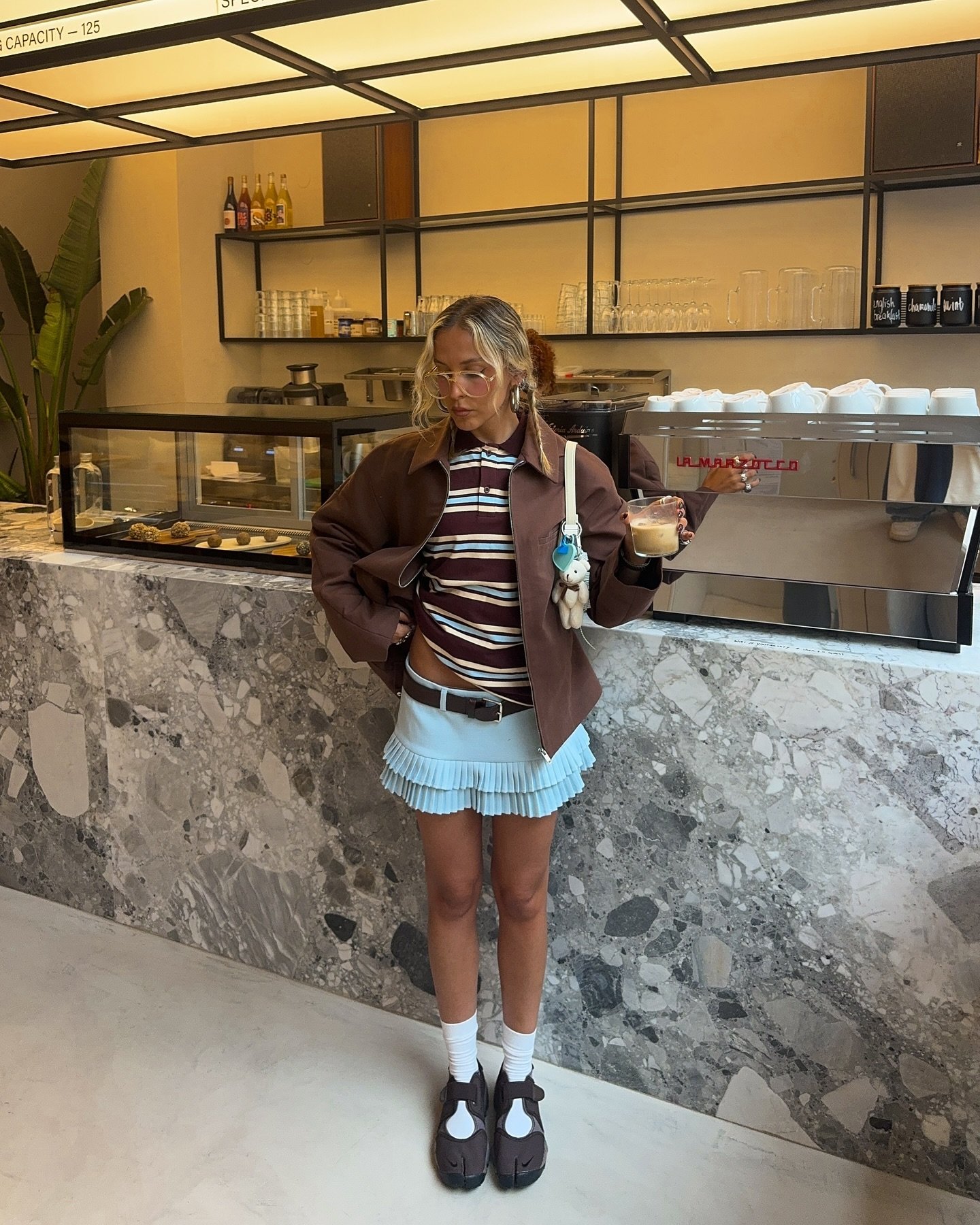 Pessoa de pé em uma cafeteria usando Nike Air Rift marrons, blusa listrada e saia azul clara, segurando um café.