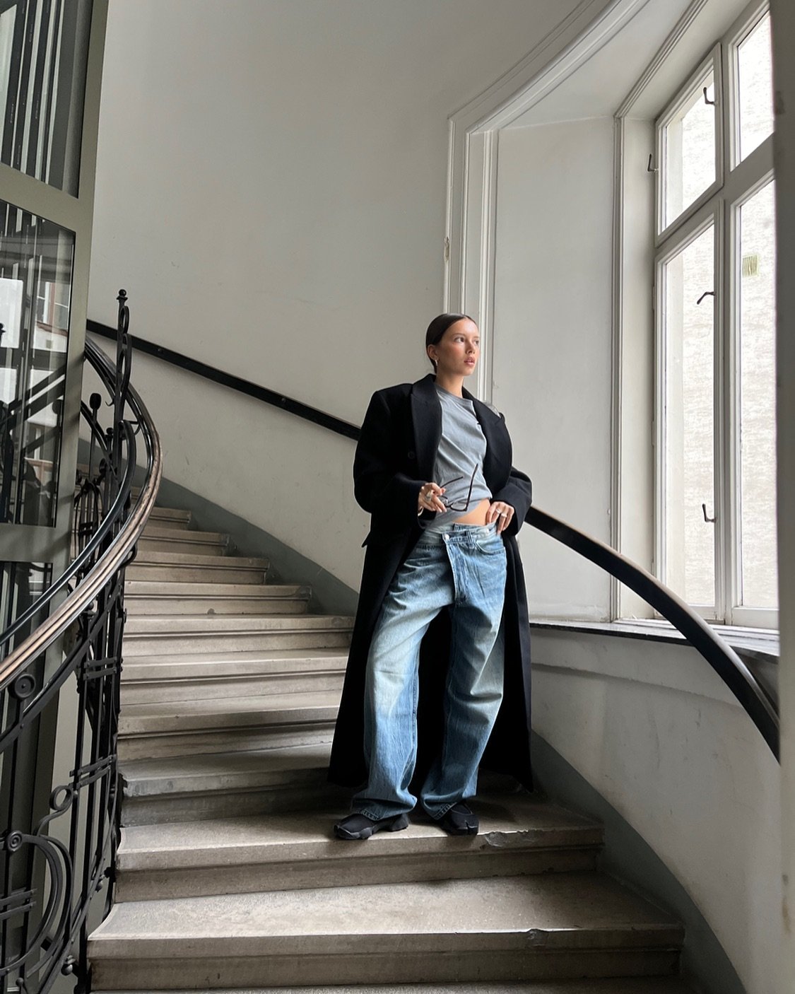 Pessoa em escada, vestindo casaco longo, calça jeans e Nike Air Rift, segurando óculos. Ambiente iluminado por janela grande.
