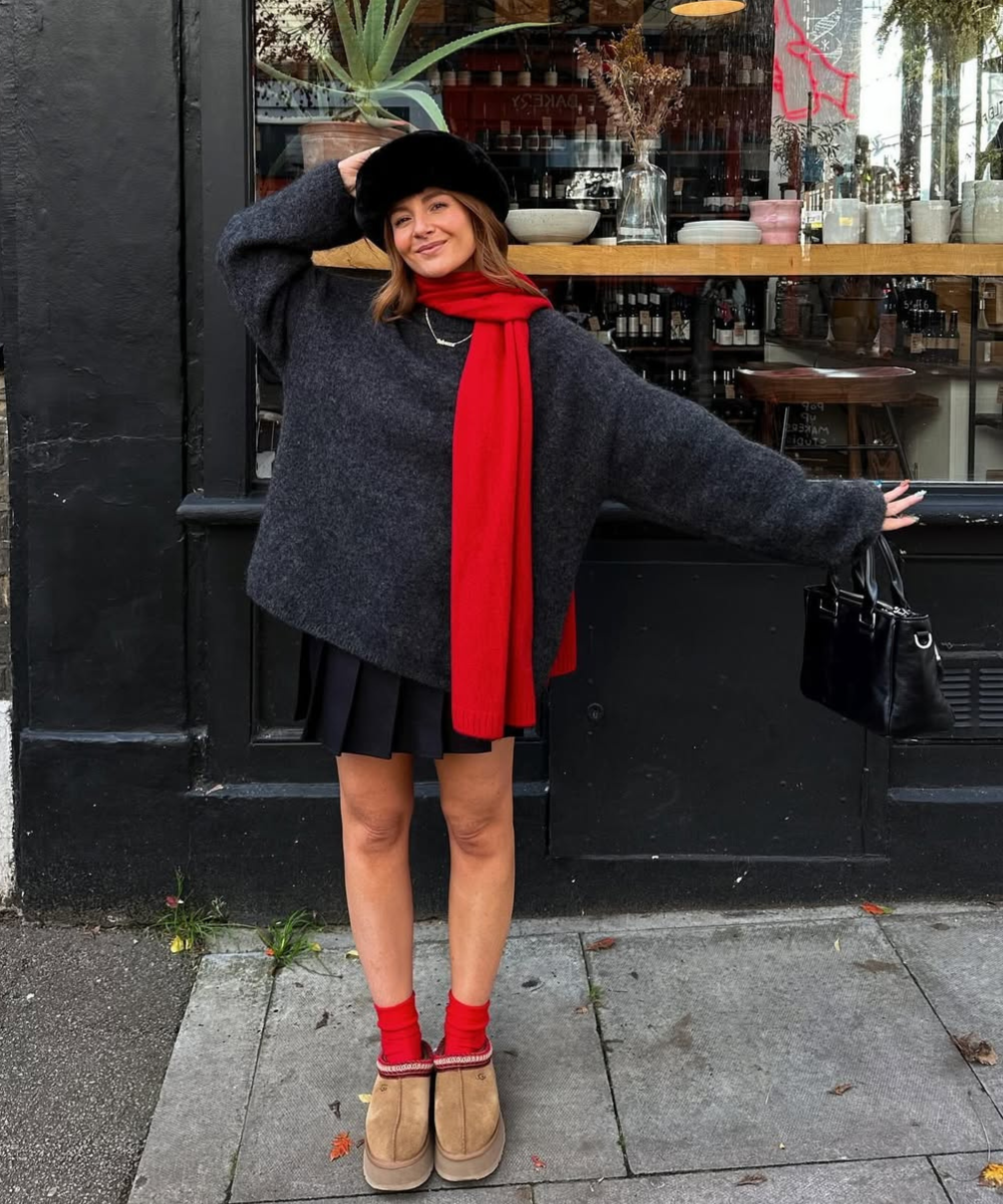 Pessoa sorrindo, vestida com suéter cinza, cachecol vermelho e chapéu preto. Usa sapato forrado marrom e segura bolsa preta.