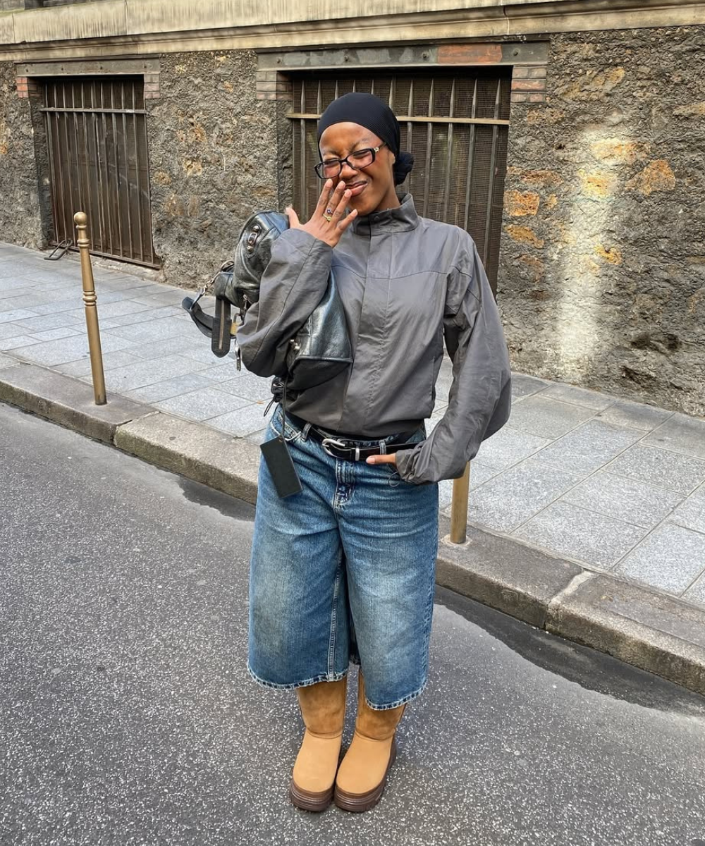 Pessoa sorridente na rua usa jaqueta cinza, bermuda jeans e sapato forrado marrom, segurando uma bolsa preta.