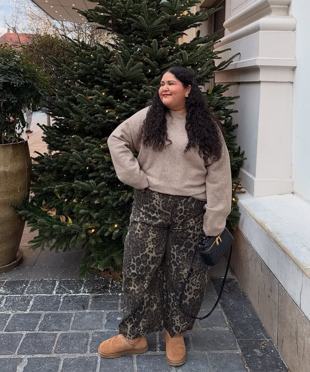 Pessoa com suéter bege, calça estampada e sapatos forrados de camurça, em frente a uma árvore natalina.