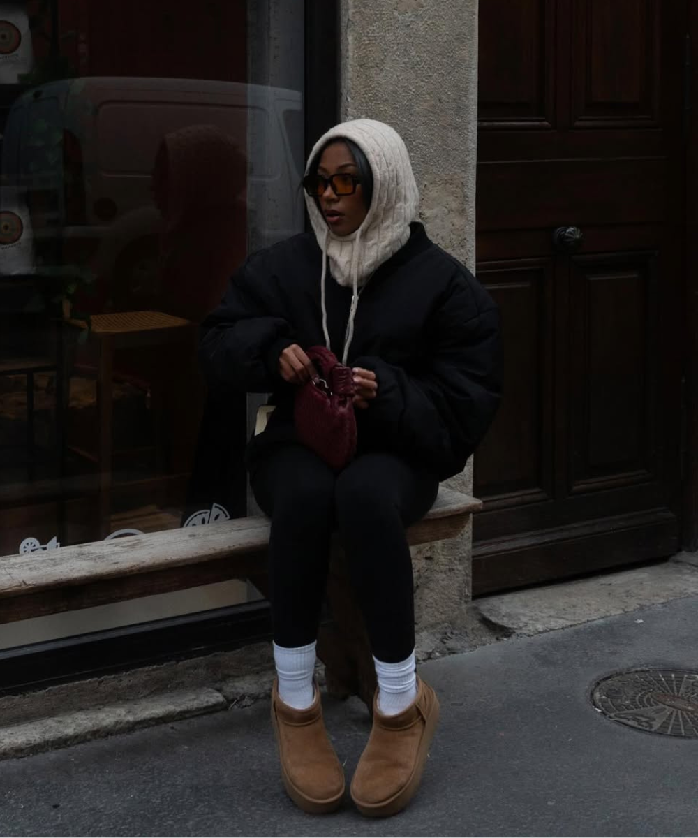 Pessoa sentada em um banco de madeira, usando casaco preto e sapato forrado marrom, segurando bolsa vermelha.