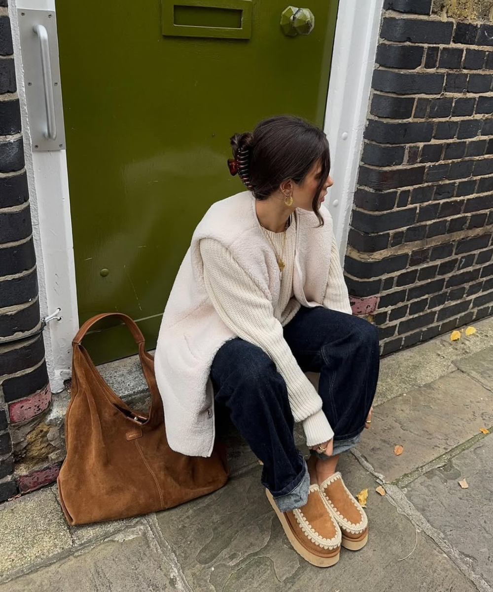Mulher sentada na calçada, usando casaco e sapato forrado. Bolsa marrom ao lado, porta verde atrás.