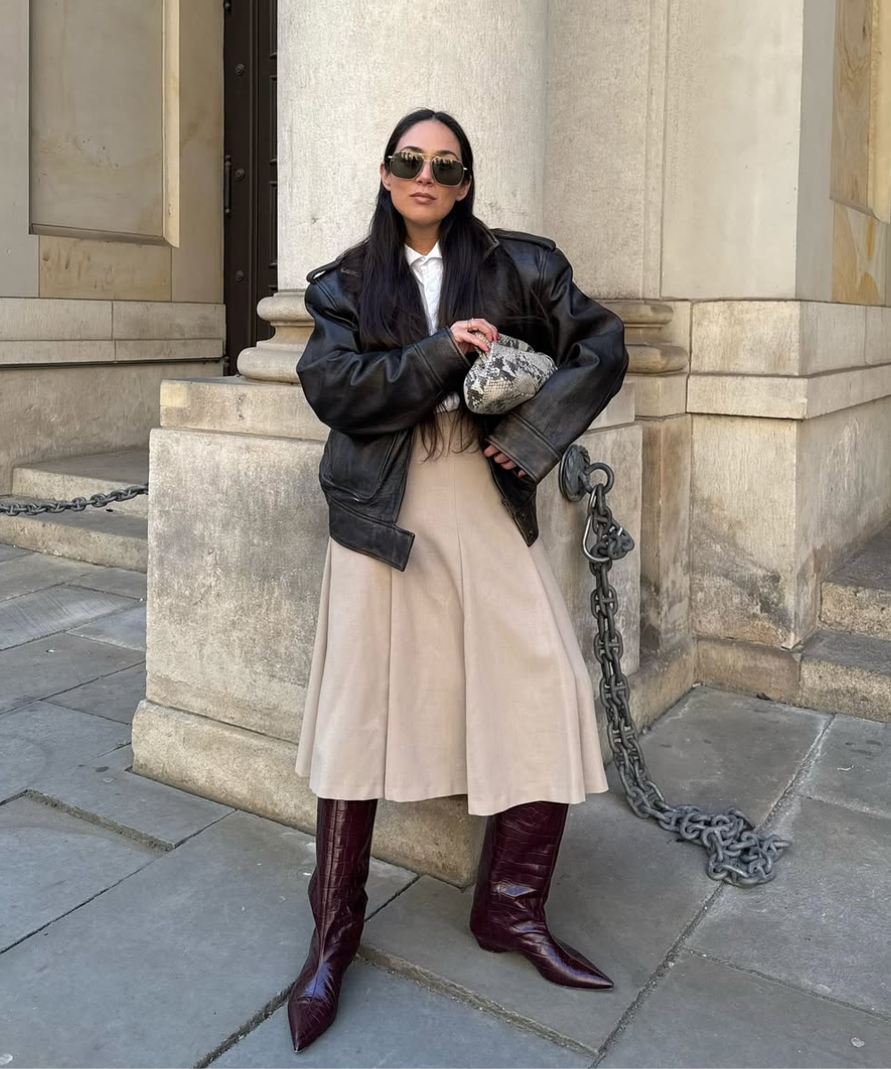 Mulher com óculos escuros usa jaqueta preta, saia bege e botas longas em looks de inverno com saia.