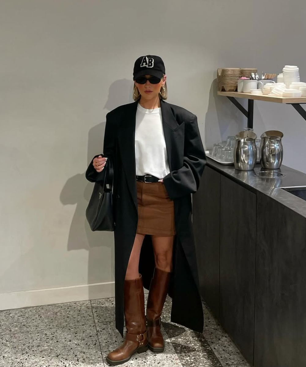 Pessoa com casaco longo, saia marrom e botas, compondo um dos looks de inverno com saia, em ambiente interno.