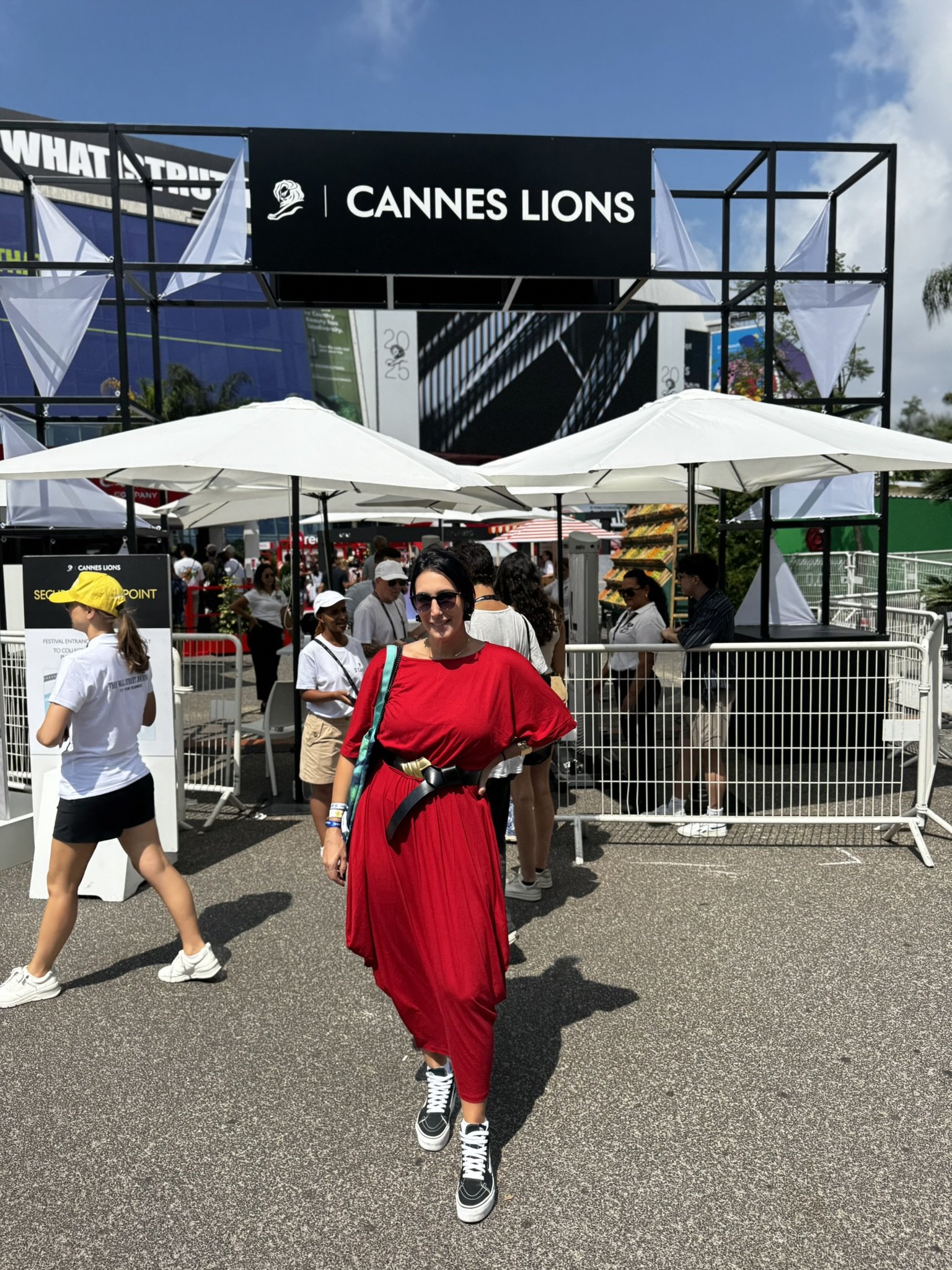 Participante em frente à entrada do Cannes Lions, usando vestido vermelho e tênis, sob sol intenso do verão europeu.
