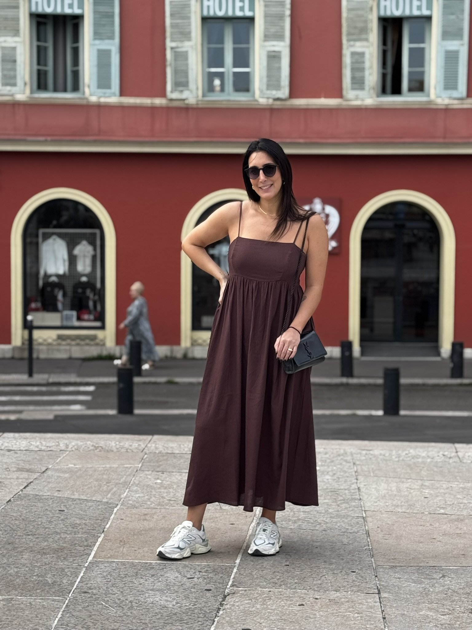 Mulher de óculos escuros em frente a prédio vermelho, vestindo um vestido marrom e tênis, representando um cenário de verão europeu.