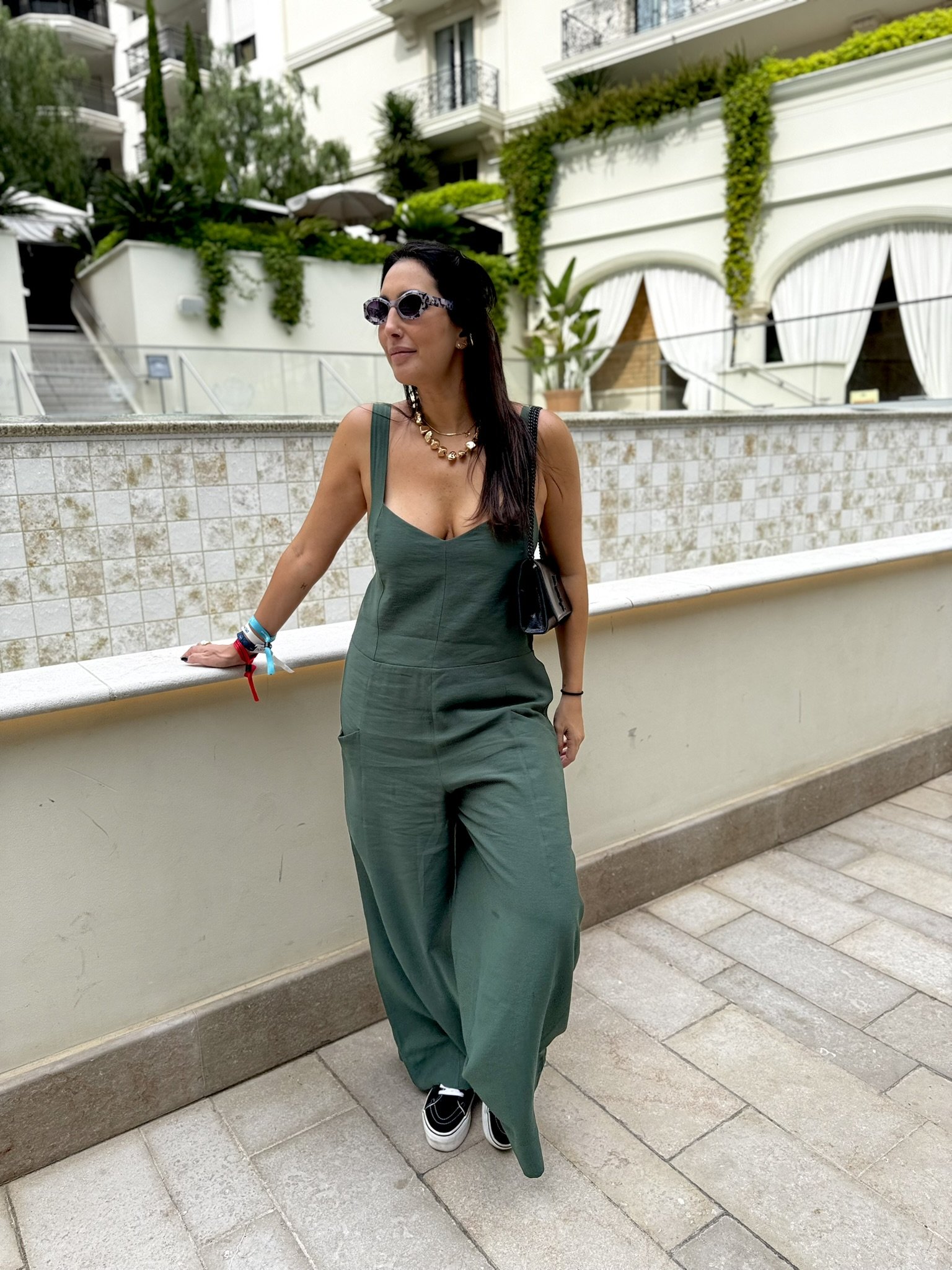 Mulher de macacão verde e óculos escuros em uma varanda, clima descontraído de verão europeu.