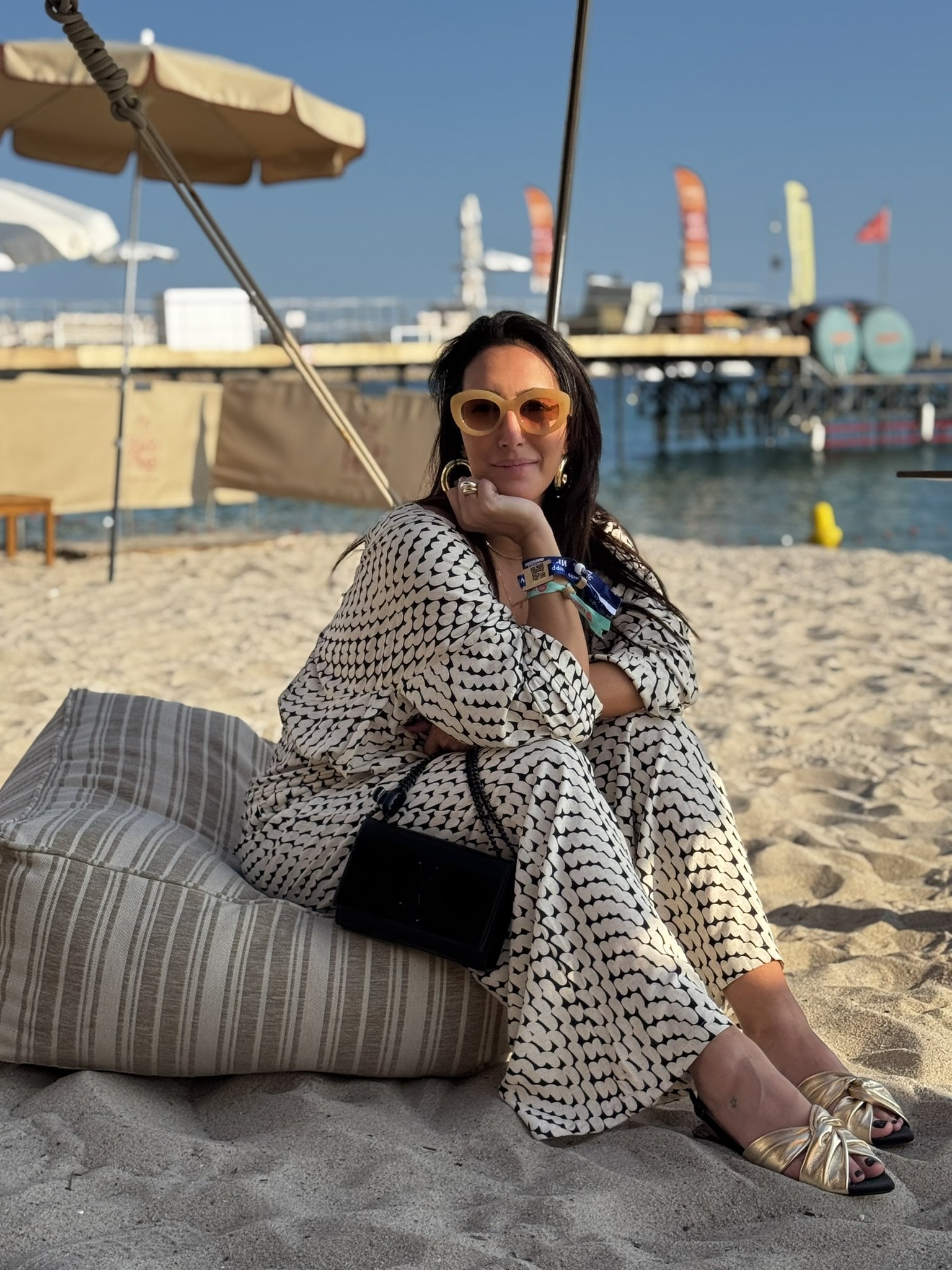 Mulher sentada em praia ensolarada, desfrutando do verão europeu, com óculos de sol e guarda-sóis ao fundo.