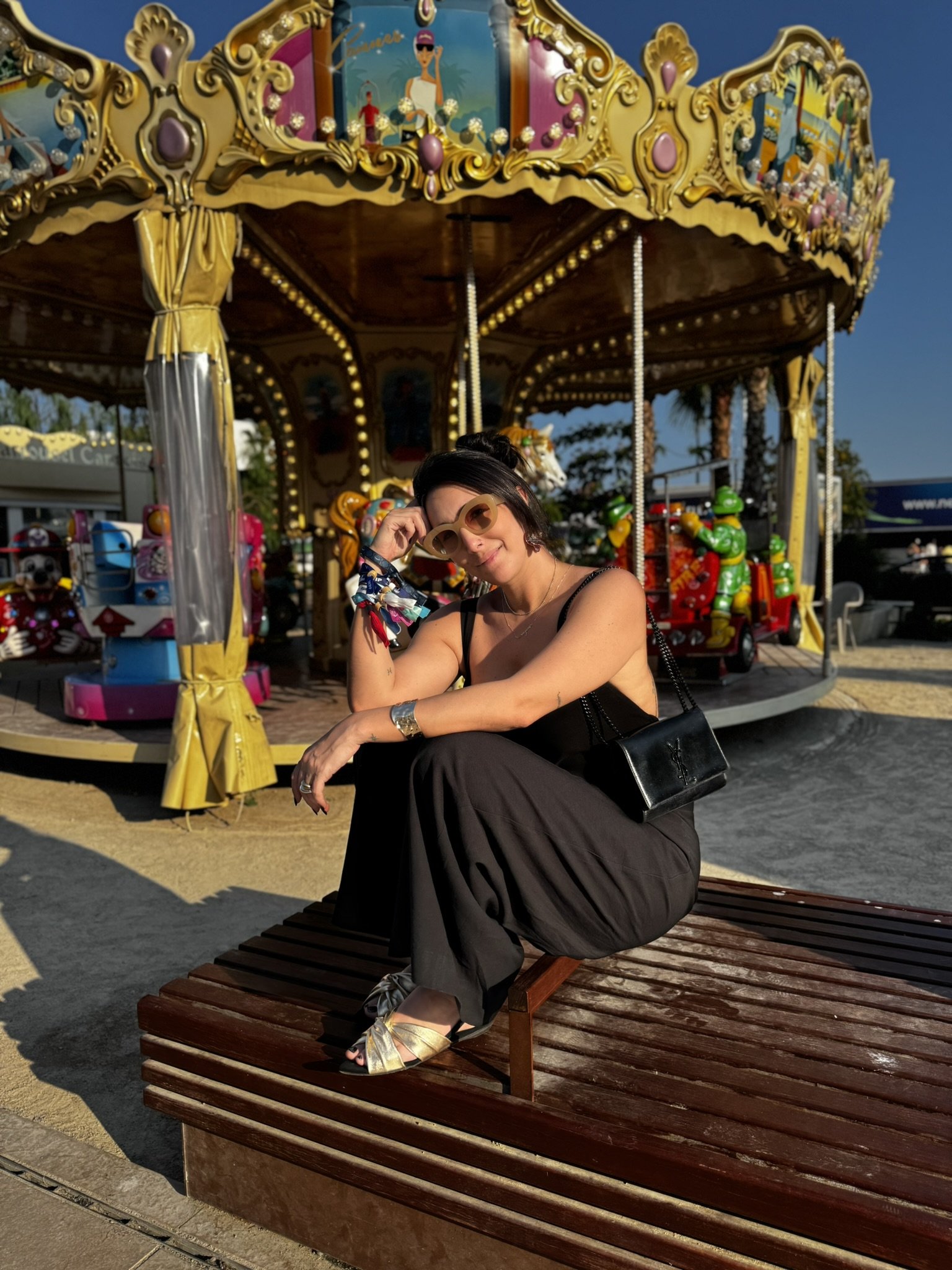 Mulher sentada em um banco de madeira próximo a um carrossel vintage durante o verão europeu, usando óculos escuros.