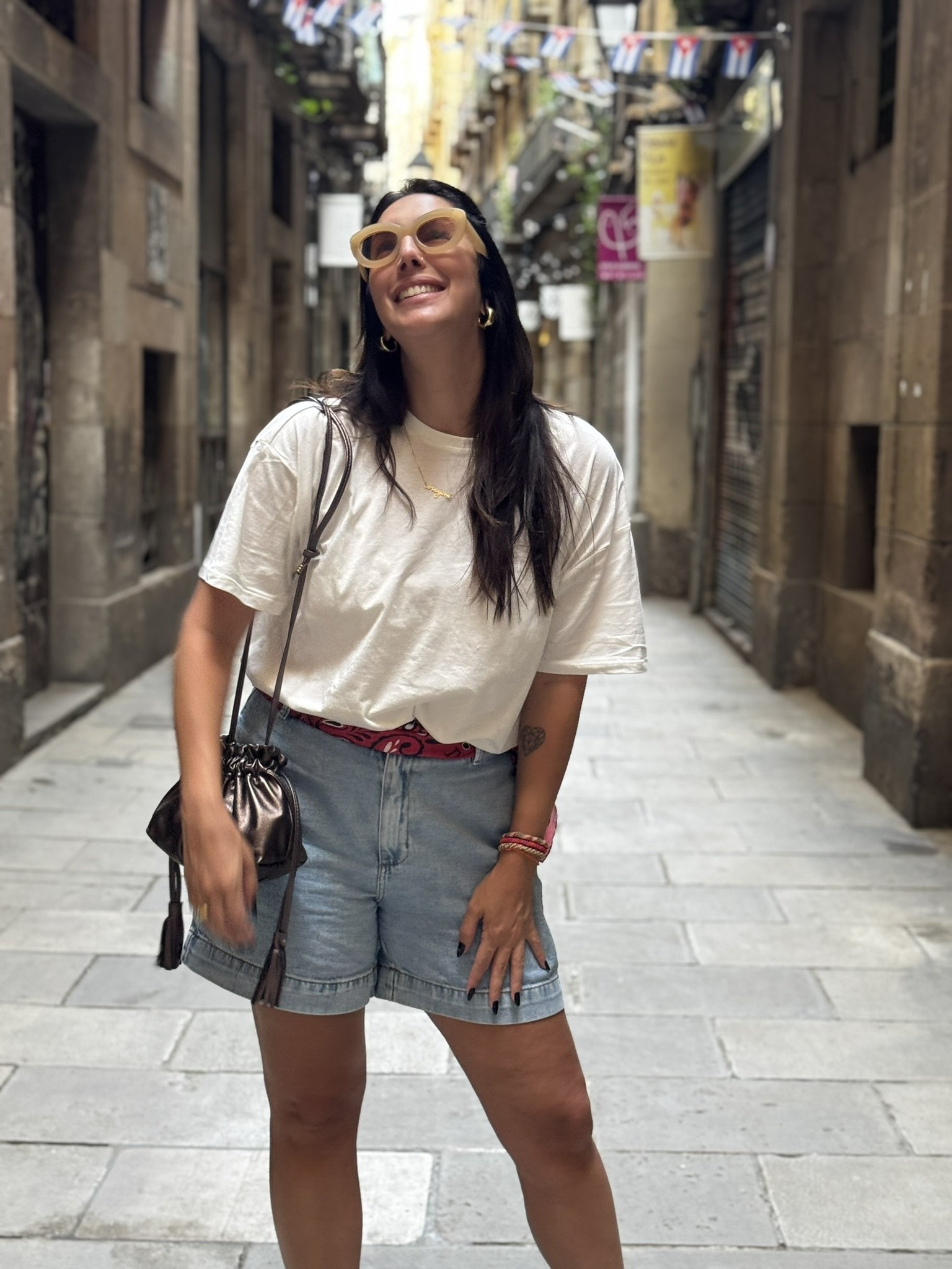 Mulher sorridente em rua europeia com decoração, usando óculos de sol, shorts e camiseta, clima de verão europeu.