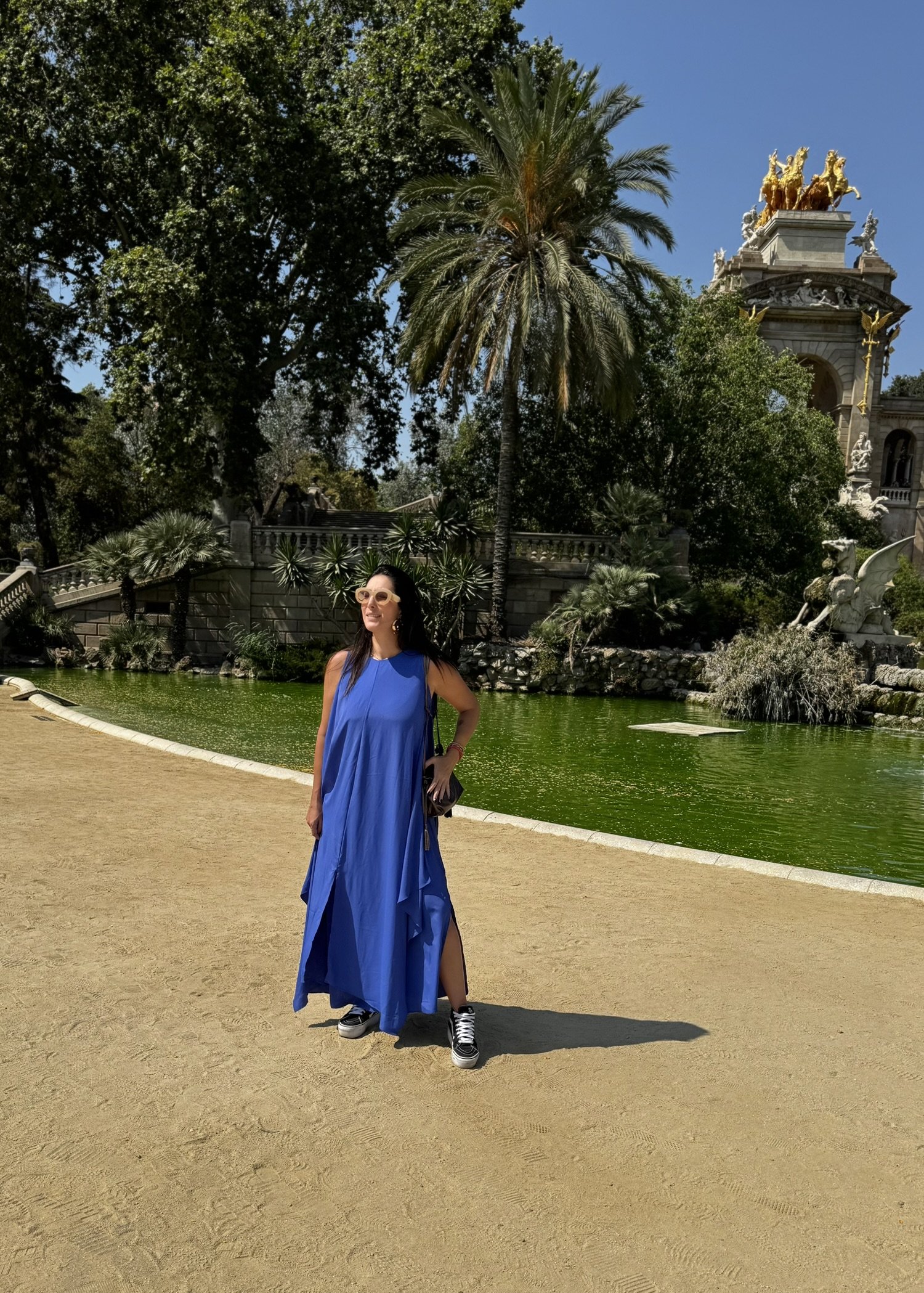Mulher de vestido azul em parque europeu com palmeiras e escultura ao fundo, sob céu claro de verão.