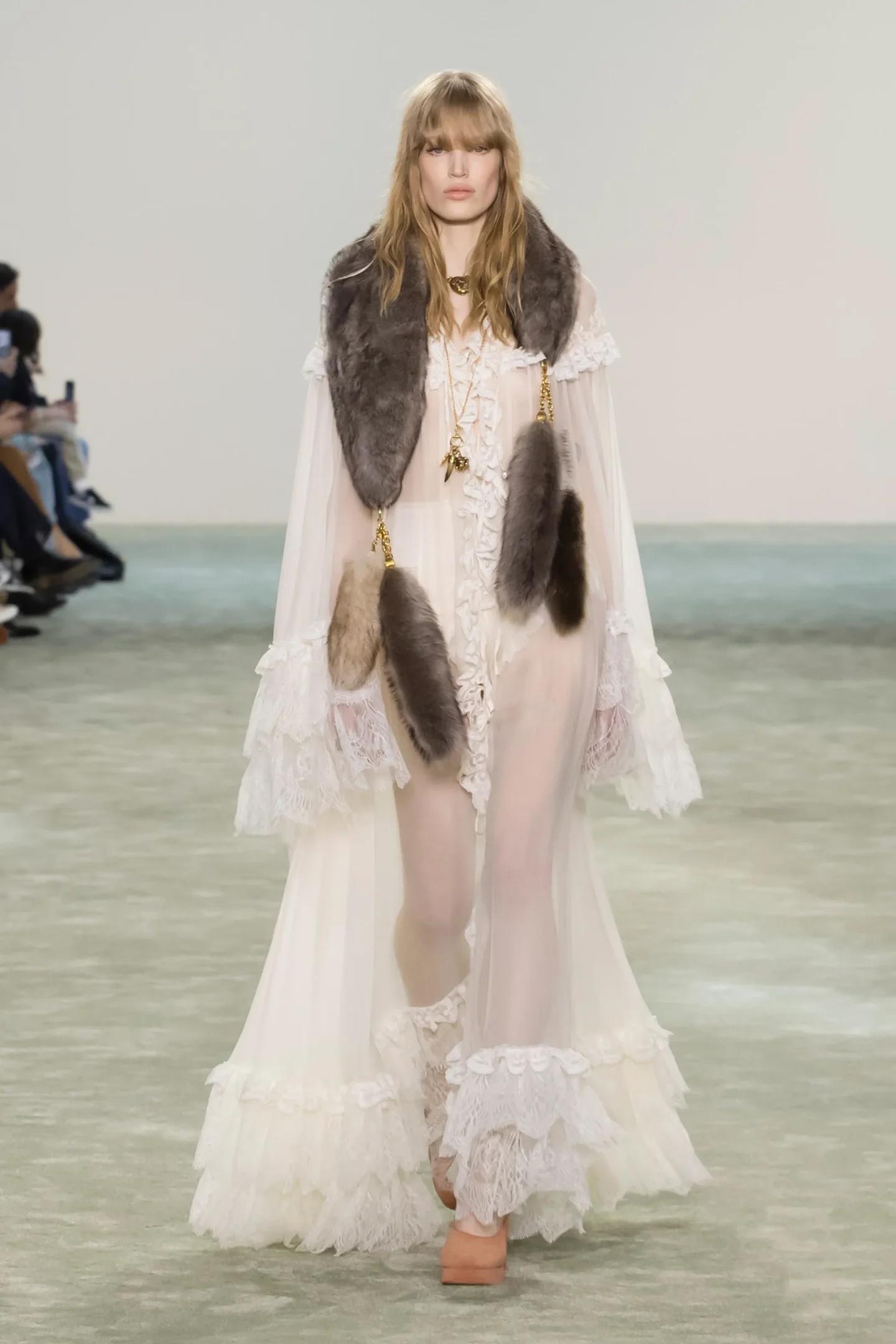 Desfile de moda apresenta modelo com vestido esvoaçante e detalhes em pelos, destacando chinelo de dedo.