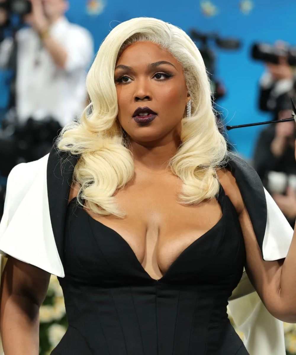 Pessoa em um evento com cabelo loiro ondulado, vestindo roupa preta elegante. Fundo com câmeras e fotógrafos.