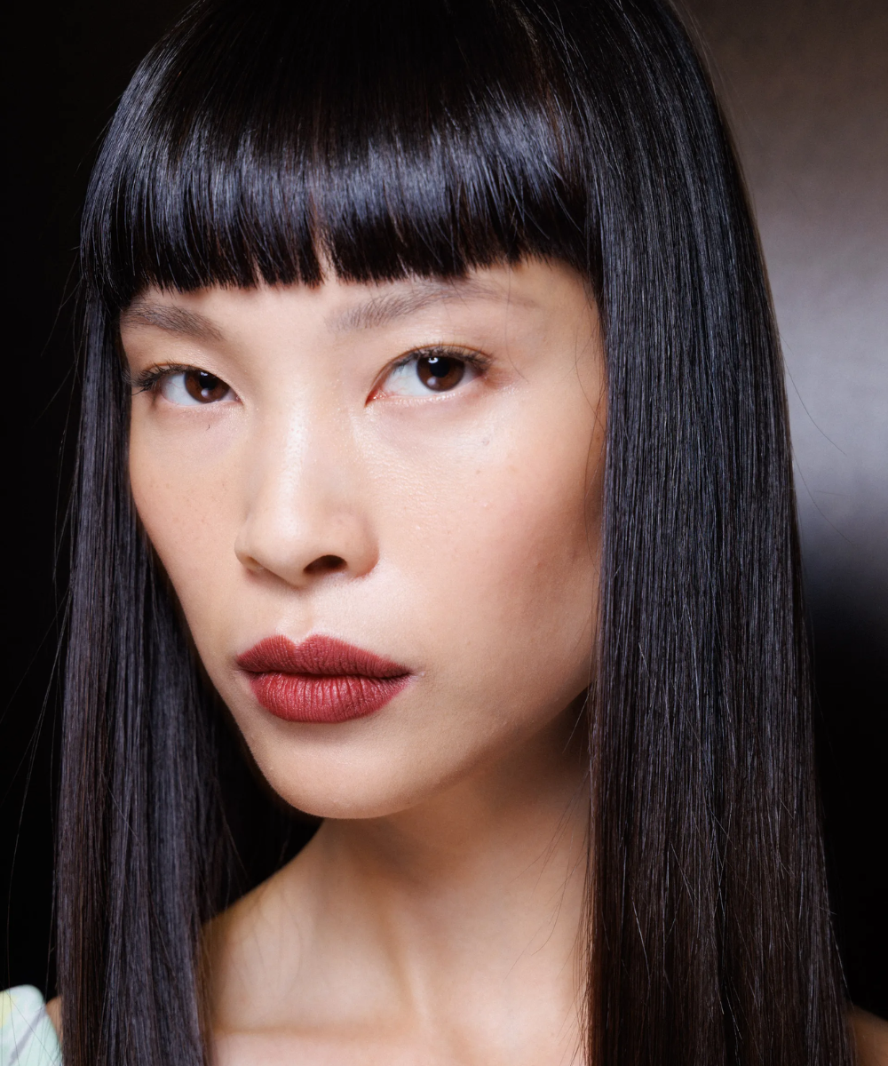 Mulher de cabelos lisos e longos com franja reta, maquiagem discreta e batom vermelho, destacando técnica de contorno.