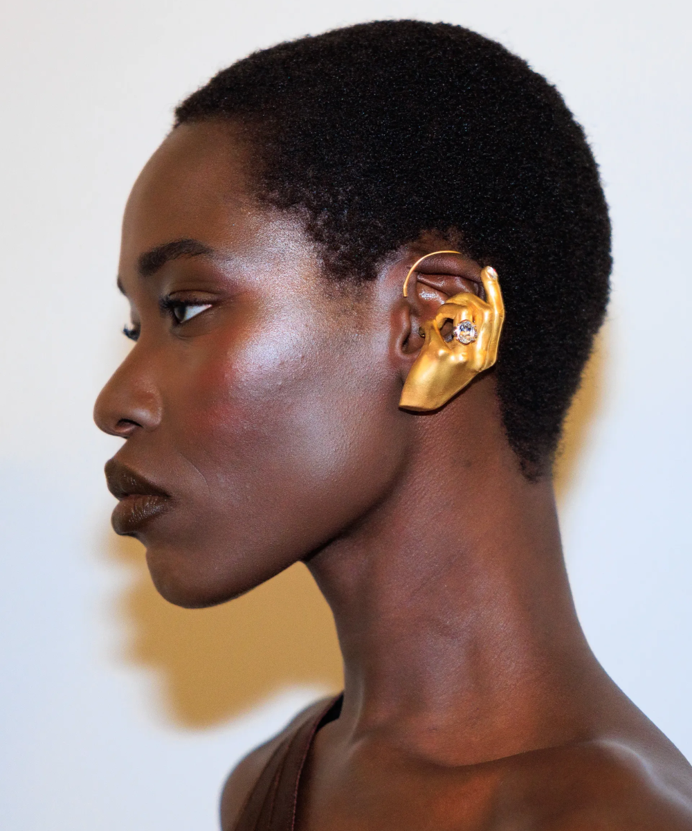 Pessoa com adorno de orelha dourado em forma de mão, destacando-se contra fundo neutro.