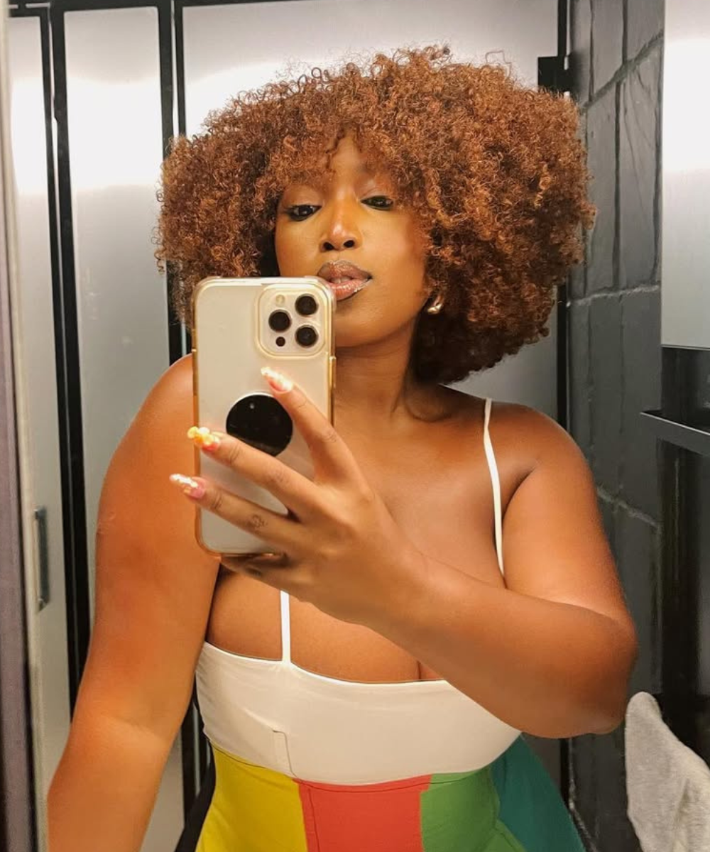 Mulher de cabelo crespo tira selfie em espelho de banheiro depois de usar o melhor óleo para cabelo crespo.