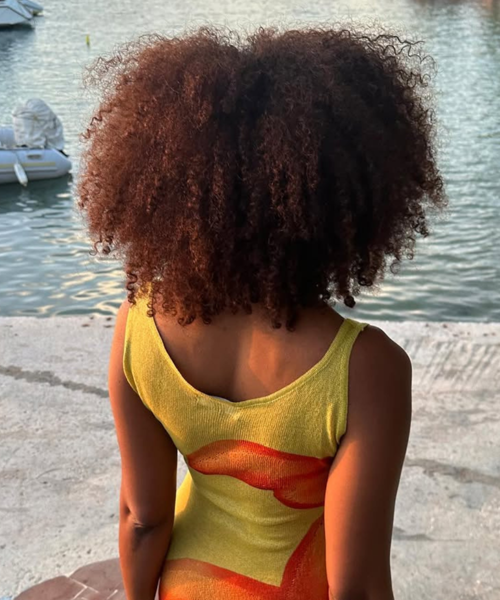 Cabelo volumoso ao ar livre, sugerindo uso do melhor óleo para cabelo crespo com brilho e definição. Fundo de lago com barco.