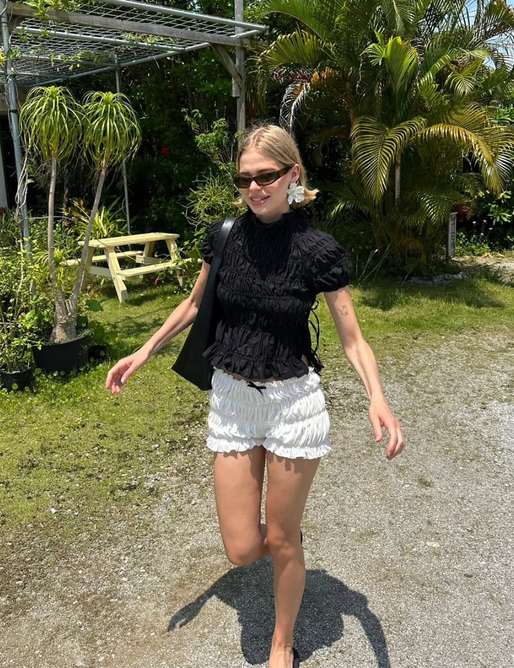 Pessoa caminhando em jardim tropical com roupas leves e óculos escuros. Verão 2026 ao fundo.