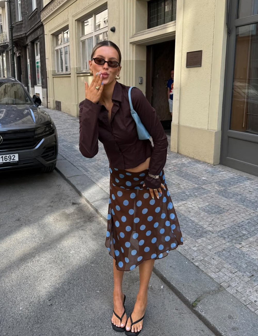 Pessoa elegante envia um beijo na calçada, com blusa escura e saia de bolinhas, em cenário urbano de Verão 2026.
