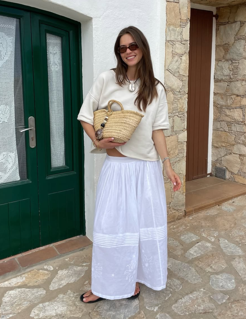 Mulher sorridente com óculos de sol e bolsa de palha, usando saia leve. Estilo casual de verão 2026.