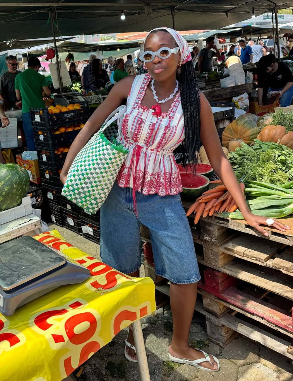 Mulher em feira ao ar livre com roupas coloridas, cercada por frutas e verduras. Verão 2026.