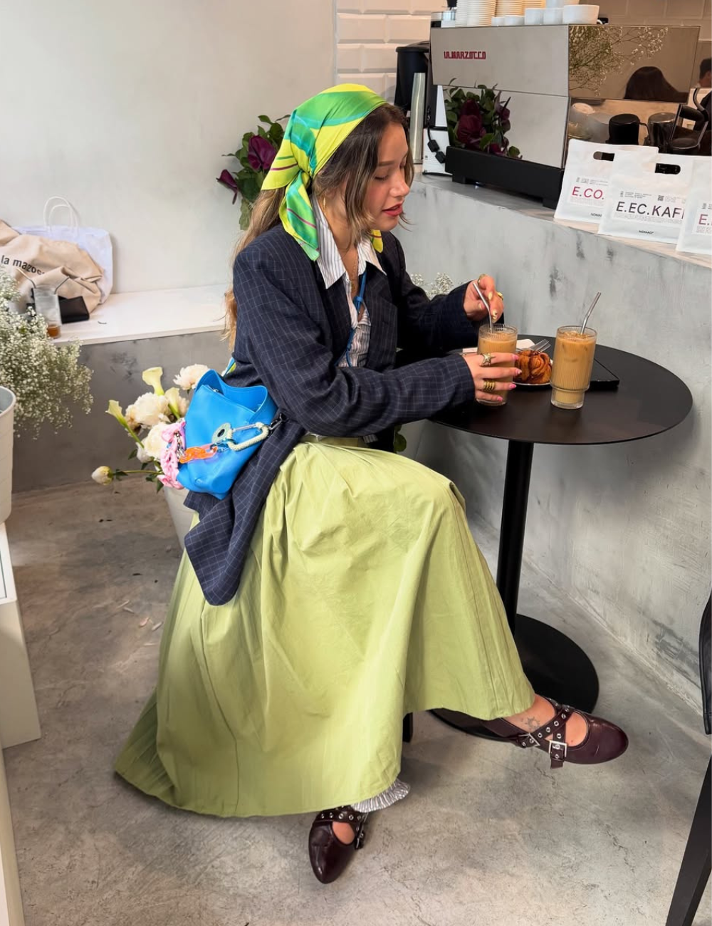 Pessoa elegante em café, com lenço verde e saia, aproveita bebida refrescante. Estilo tendência Verão 2026.