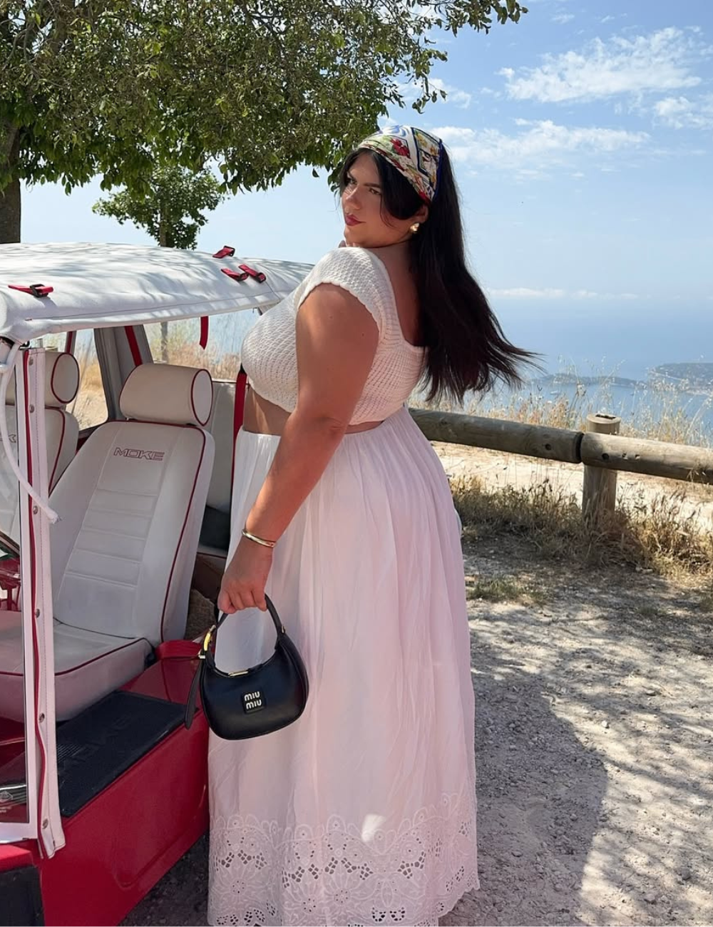 Pessoa com vestido branco, bolsa preta e lenço, posando ao lado de bugue vermelho em cenário ensolarado. Perfeita para o Verão 2026.