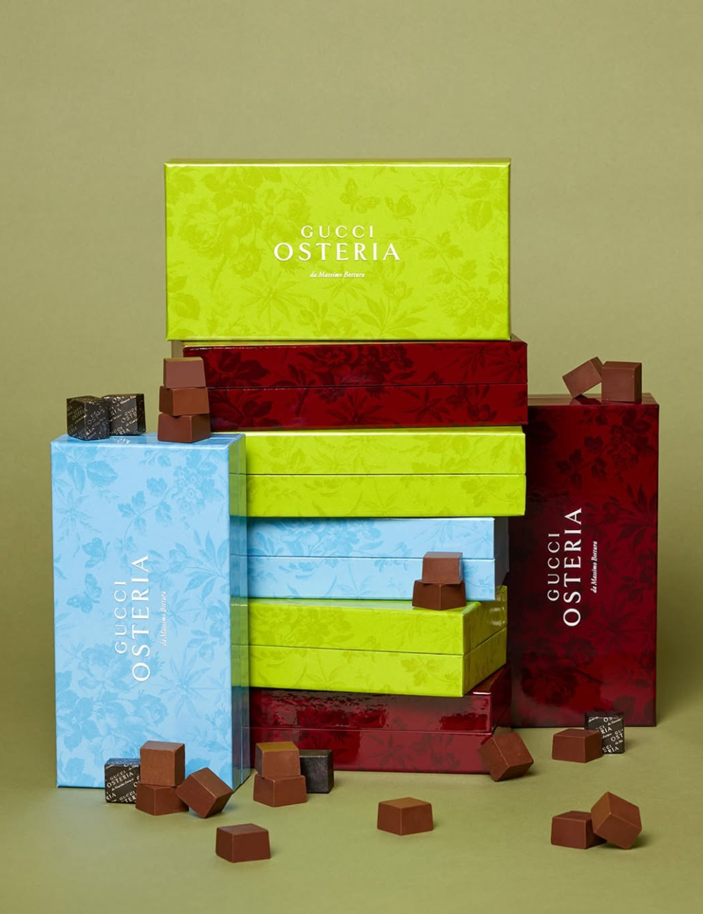 Caixas elegantes da Gucci Osteria empilhadas, cercadas por chocolates gourmet. Toque de moda na apresentação da comida.