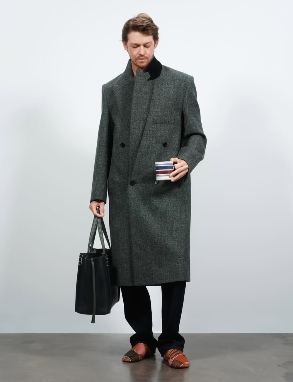 Homem com casaco elegante segura bolsa e copo listrado, destacando moda contemporânea.