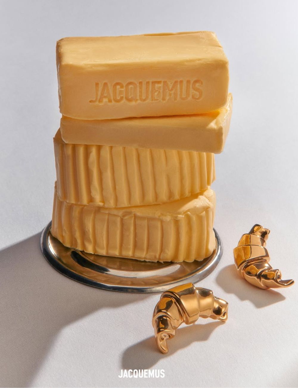Pedaços de manteiga com o nome Jacquemus empilhados sobre um prato, ao lado de acessórios dourados em forma de croissant.