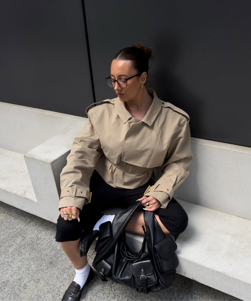 Mulher sentada em banco com trench coat bege e calças pretas, segurando bolsa preta. Calçado e acessórios complementam o visual.