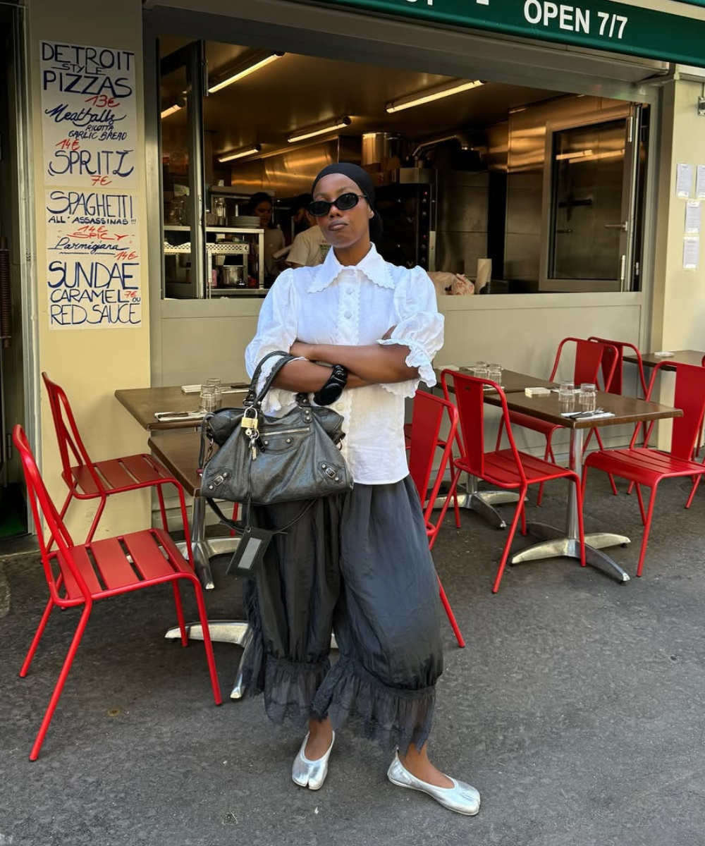 Pessoa em frente a restaurante veste camisa branca e calças largas tendência, segurando bolsa.