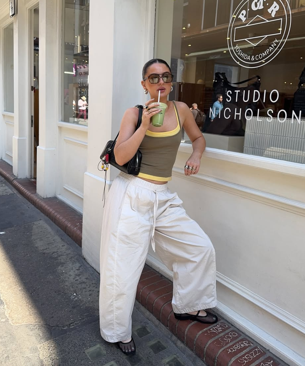 Pessoa usando calça larga branca e regata, bebendo smoothie em frente a uma loja. Modelos de calças tendência.