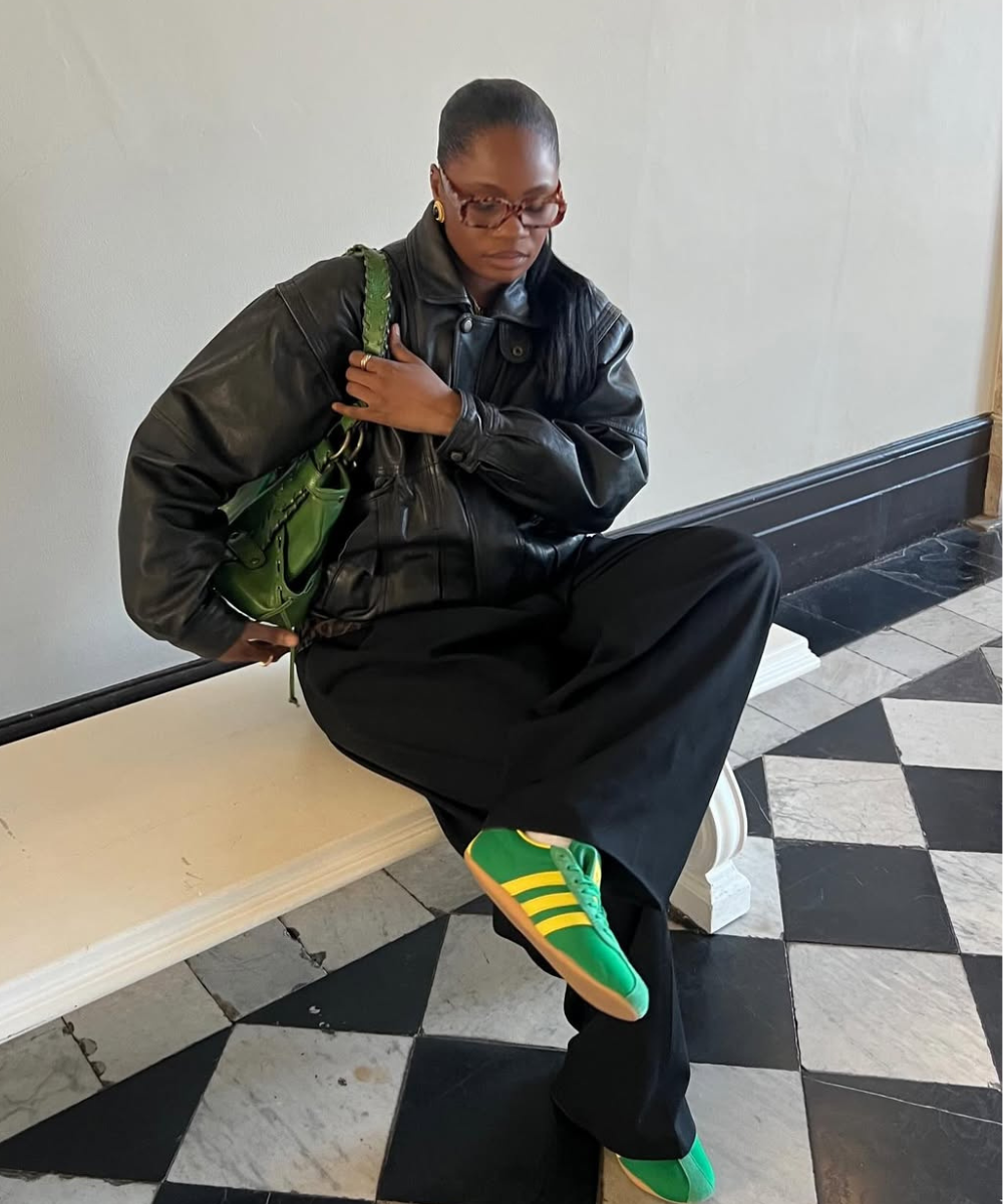 Pessoa com jaqueta de couro preta e calças largas, sentada em banco. Sapatos verdes e bolsa combinam. Modelos de calças tendência.