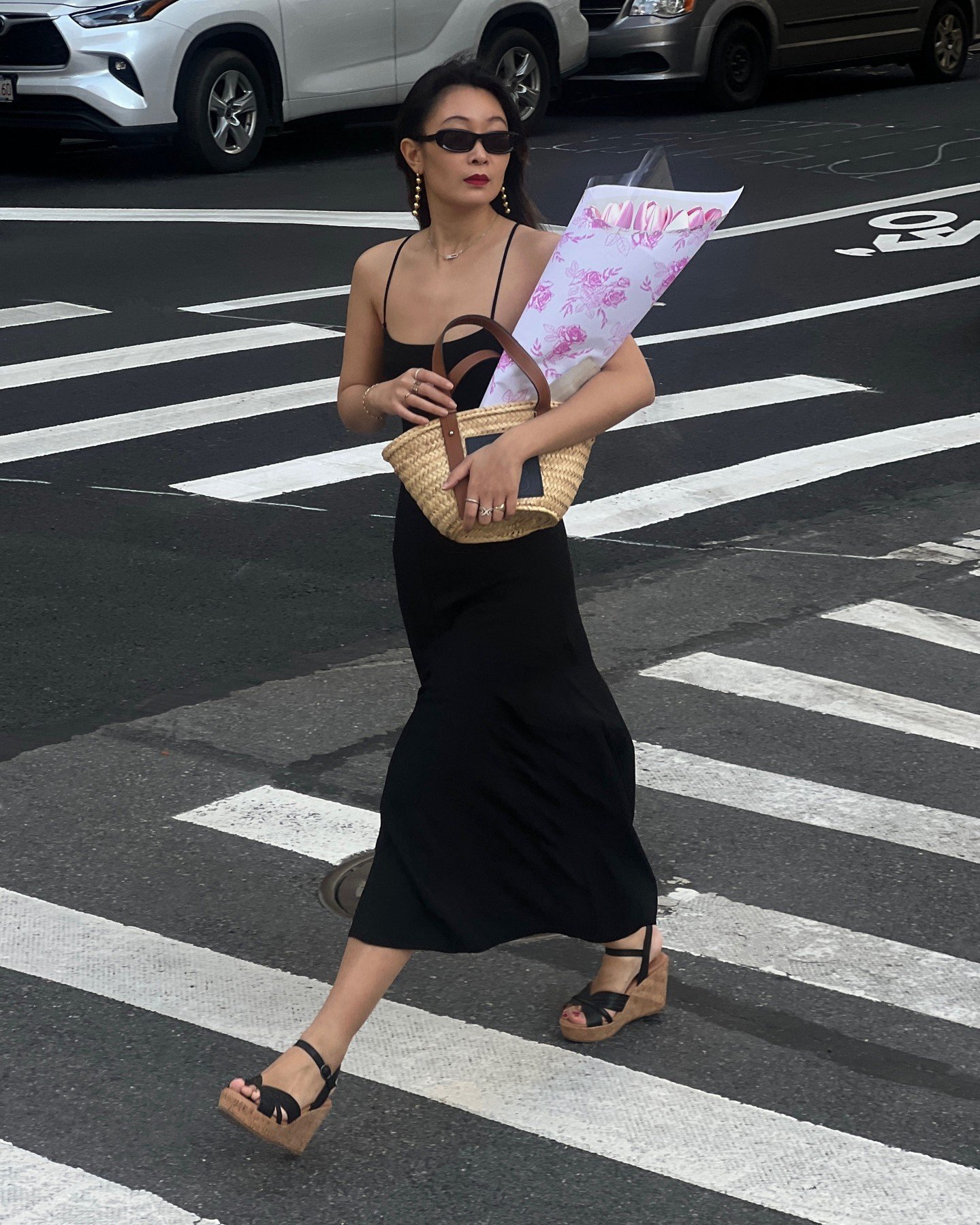 Pessoa atravessa rua usando vestido preto e sandálias plataforma. Segura flores e bolsa de palha.