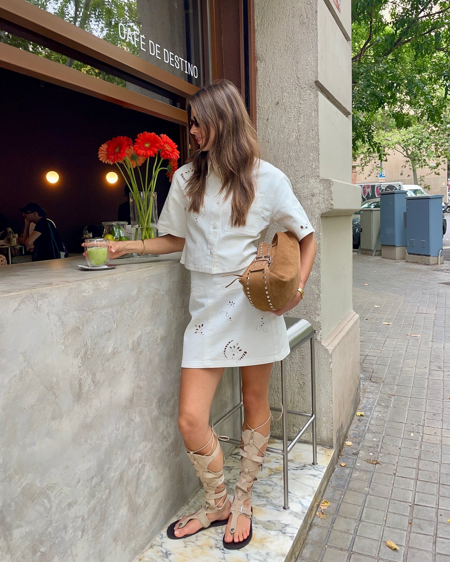 Pessoa usando sandálias gladiadoras bege e roupa branca em frente a um café, segurando uma bolsa e um copo de bebida.