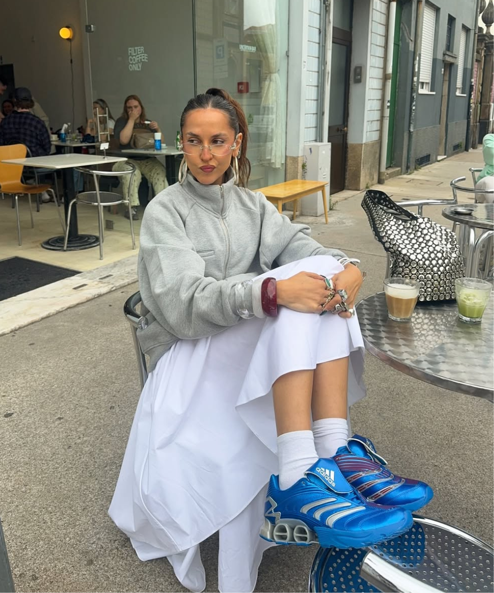 Mulher sentada de moletom cinza com gola zíper, saia branca, tênis azuis, em uma mesa de café ao ar livre.
