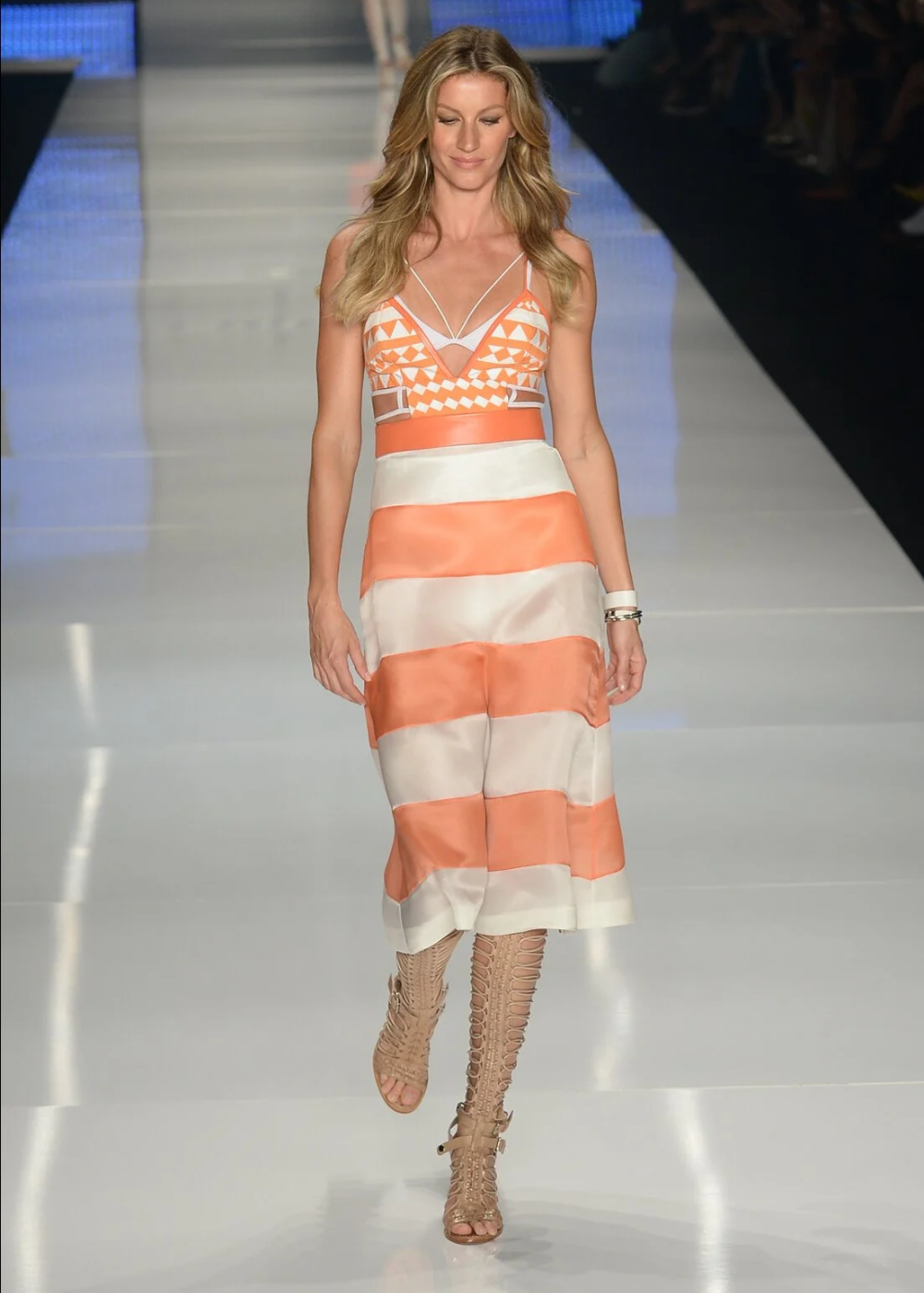 Modelo desfilando em passarela usando vestido laranja e bege com sandálias gladiadoras.