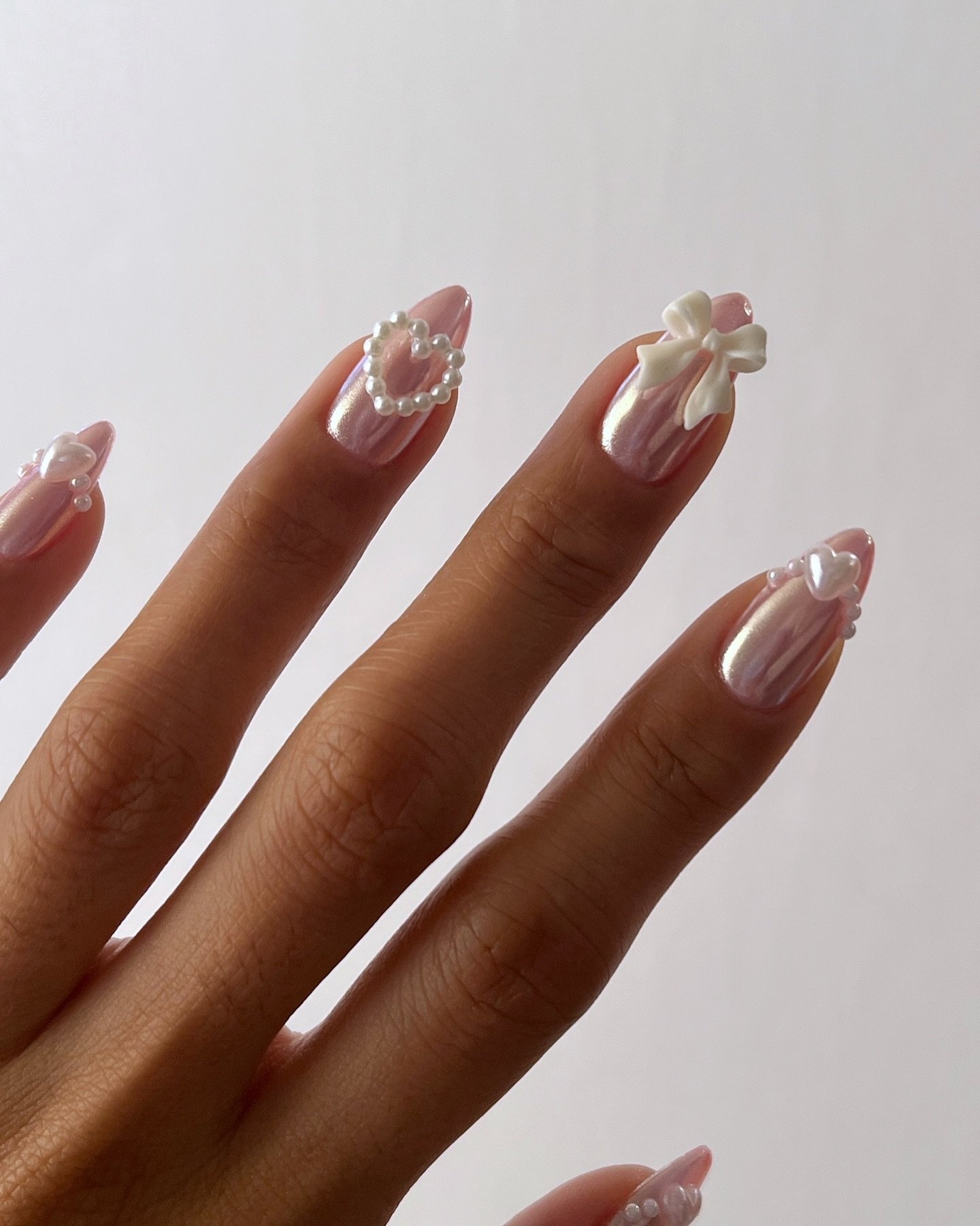 Unhas com esmalte rosa perolado e decoração de pérolas, laço e corações, exibindo nail arts estilosas.