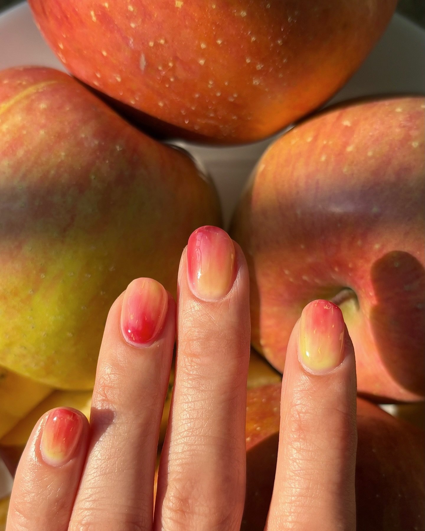 Unhas com degradê de cores semelhantes a maçãs frescas ao fundo, representando nail arts estilosas.