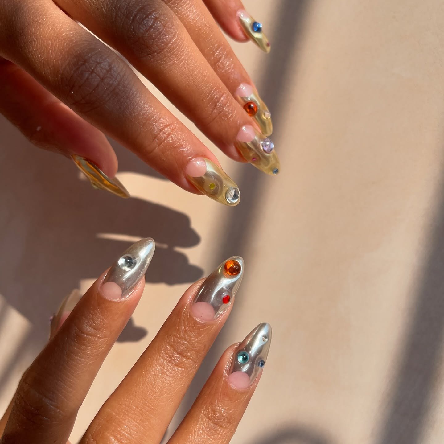 Unhas decoradas com esmalte metálico e pedras coloridas, representando nail arts estilosas.