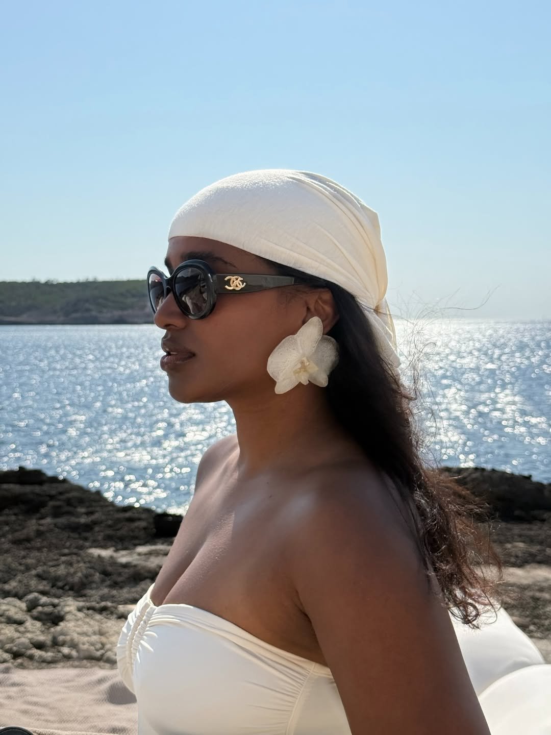 Pessoa de lenço e brincos grandes, usando óculos de sol do momento, diante de um mar brilhante.